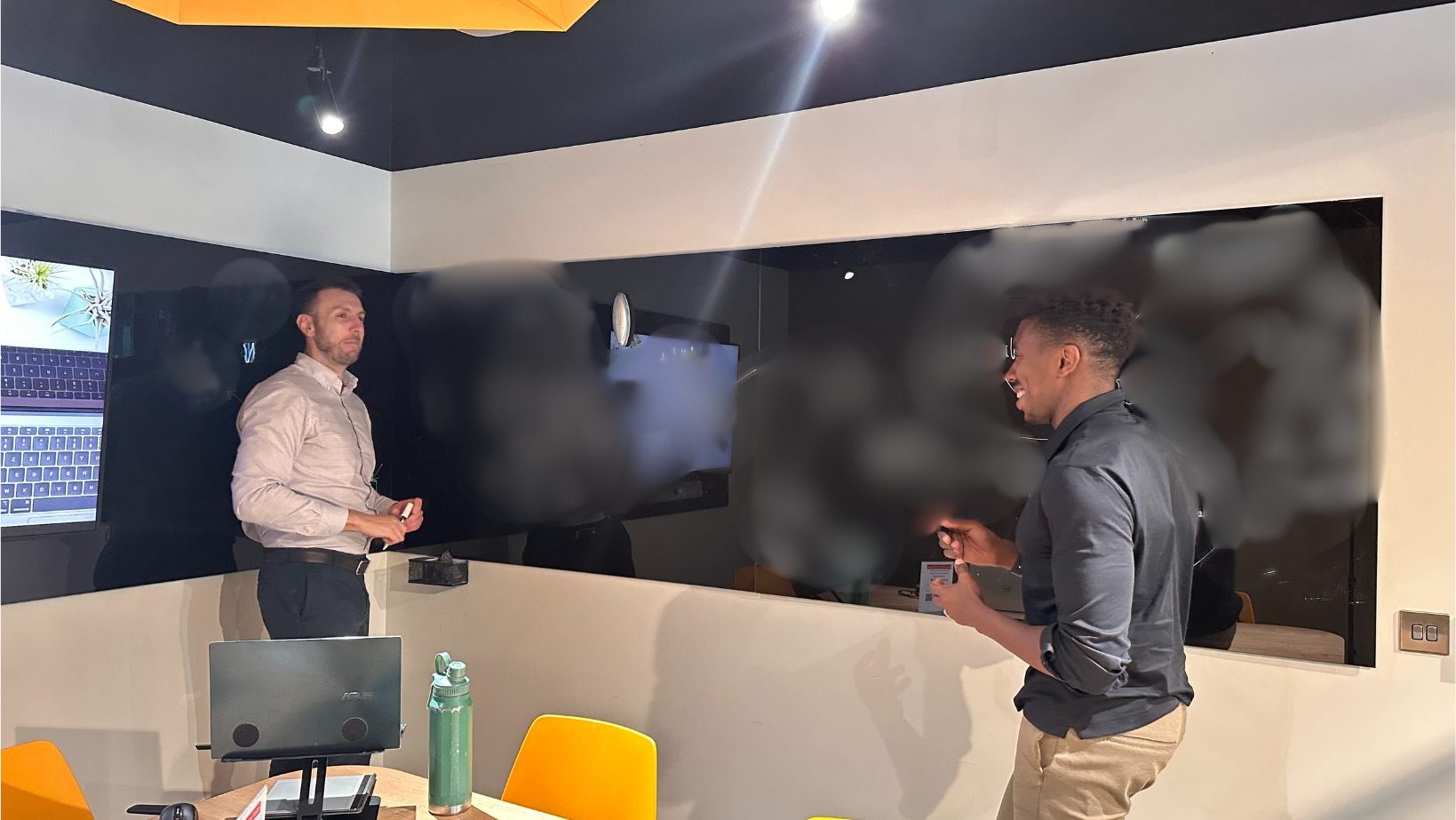 John and a client at a glass-wall whiteboard, mid-session, board content obscured for confidentiality