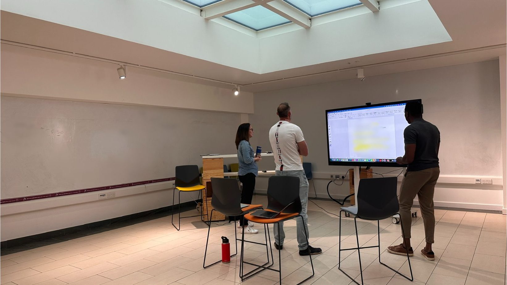 A small group session in a skylit workshop room with whiteboard wall and shared screen
